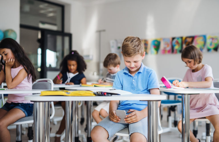 A small school boy with smartphone sitting at the desk in classroom, playing.